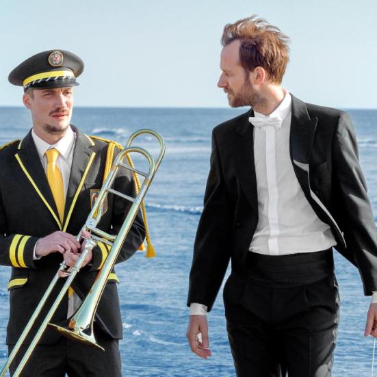 To mand der står ved havet iført sort tøj og hvide skjorter den ene har et blæser instrument i hænderne