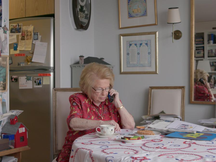 En ældre kvinde der sidder med en telefon mod sit øre ved et bord i et køkken