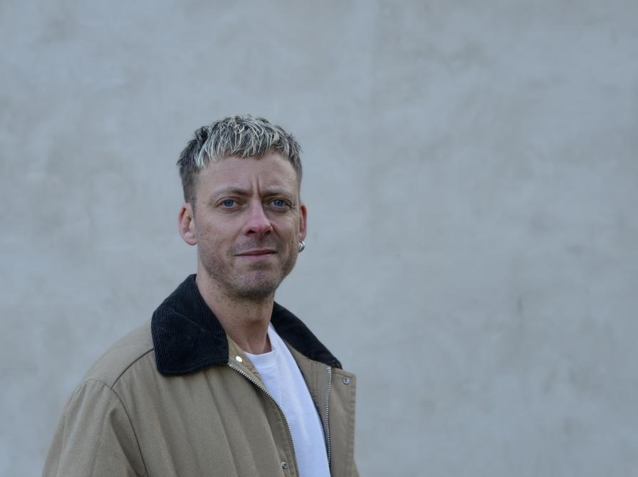 Mand der står foran en grå murstensvæg og ser lige ind i kammaret med en jakke på