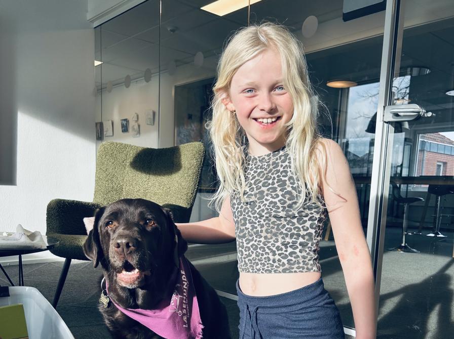 en mørkebrun labrador poserer sammen med en lyshåret pige. der står en kasse med bøger og hunden har et lilla læsehundstørklæde om halsen